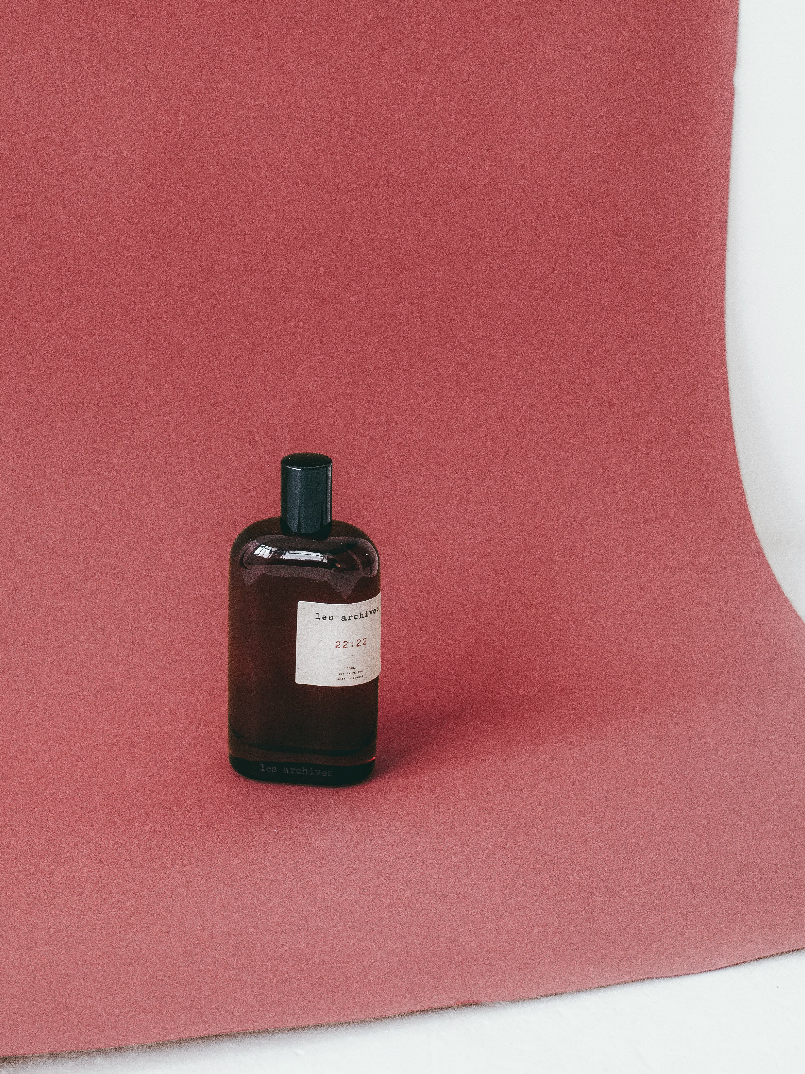 Flacon de parfum Les Archives 22:22 en verre fumé, posé sur un fond rouge texturé, photographie minimaliste en studio.