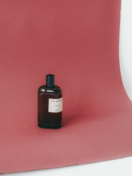 Flacon de parfum Les Archives 22:22 en verre fumé, posé sur un fond rouge texturé, photographie minimaliste en studio.